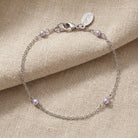 A delicate silver chain bracelet with four small light gray pearls spaced along the chain, displayed on a textured beige fabric background. The clasp is a lobster clasp with an engraved tag.