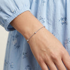 A delicate silver chain bracelet with small bead accents worn on a person's wrist, shown against a light blue floral dress.