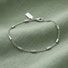 A silver chain bracelet with cable links and a lobster clasp, displayed on a textured sage green fabric background.