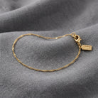 A delicate gold chain bracelet with a lobster clasp displayed on a textured gray fabric background.