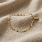 A delicate gold chain bracelet displayed on a textured beige linen fabric background.