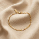 A delicate gold chain bracelet with a arrow-link design and a lobster clasp, displayed on a draped beige fabric background. 