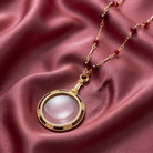Gold necklace with red beads and a magnifying glass pendant on a pink fabric background