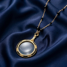 Gold necklace with black beads and a magnifying glass pendant on a blue fabric background