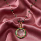 Gold necklace with green beads and a magnifying glass pendant on a pink fabric background