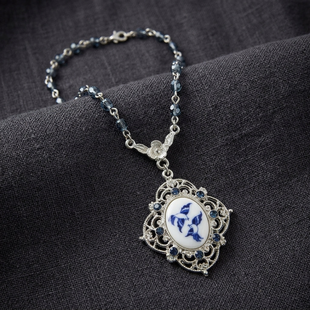 Blue beaded necklace with a Two Bird Blue Willow pendant and sapphire blue crystal accents resting on dark linen fabric surface