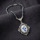 Blue beaded necklace with a Two Bird Blue Willow pendant and sapphire blue crystal accents resting on dark linen fabric surface