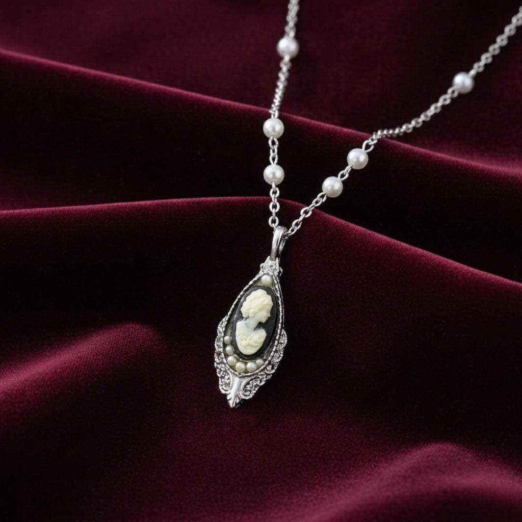 Silver tone chain necklace with a black cameo pendant and white faux pearls on the chain resting on dark red fabric