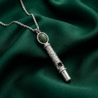 Oval Jade Gemstone Filigree Whistle Pendant Necklace resting on dark green fabric surface