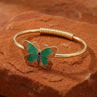 Green butterfly bracelet on a gold tone spring hinge resting on red sandstone