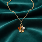 Gold necklace with a topaz crystal pendant, as well as two charms: one faux pearl and the other an amber-colored heart. All against a blue-green satin fabric background. 