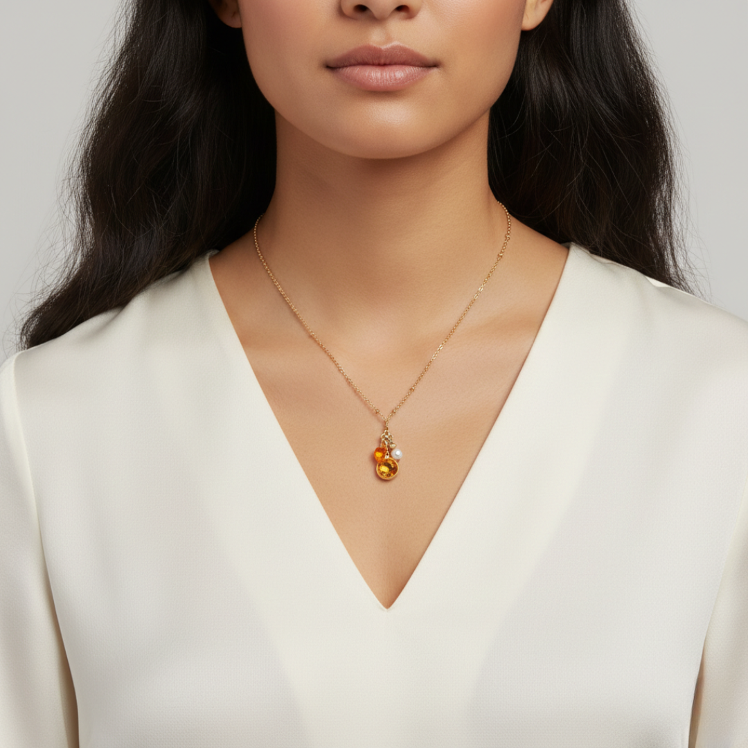 Woman wearing a necklace with a topaz crystal pendant, as well as two charms: one faux pearl and the other an amber-colored heart. All against a neutral background.