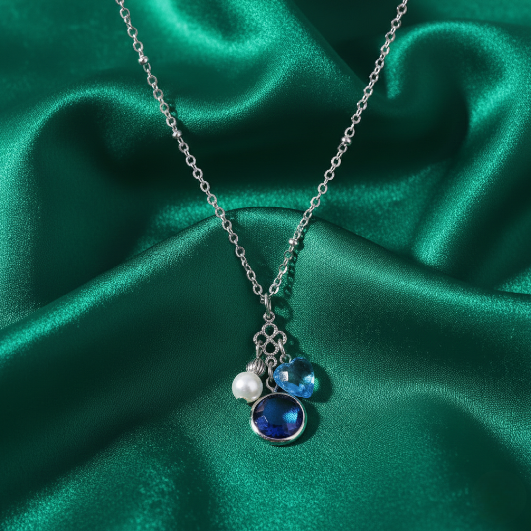 Necklace with blue crystal pendant and two charms, one of a faux pearl and the other a blue sapphire heart. All displayed on green satin fabric