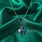 Necklace with blue crystal pendant and two charms, one of a faux pearl and the other a blue sapphire heart. All displayed on green satin fabric