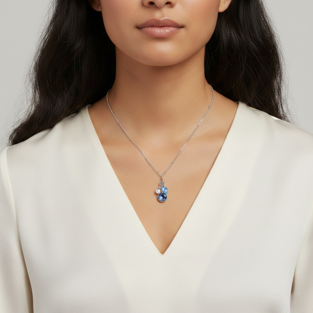 Woman wearing a necklace with a blue crystal pendant and two charms of faux pearl and blue sapphire heart against a neutral background