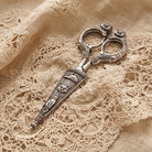 Decorative silver scissors with cat-shaped handles and embossed cat motifs on the blade, placed on beige lace fabric.