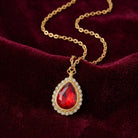 Gold tone red teardrop crystal pendant necklace with crystal accents resting on dark red fabric