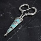Vintage-style silver tone scissors with a turquoise cover and horse designs resting on dark marble surface
