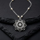 Silver tone floral pendant necklace that has clear crystal accents and white faux pearls on the chain resting on black stone surface
