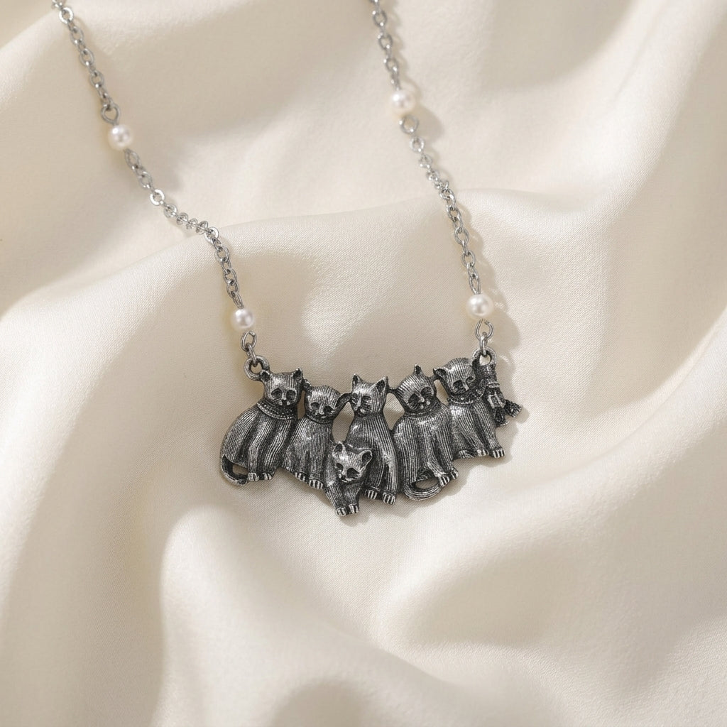Silver tone six cat pendant with white faux pearl accents on the chain resting on white colored satin fabric 