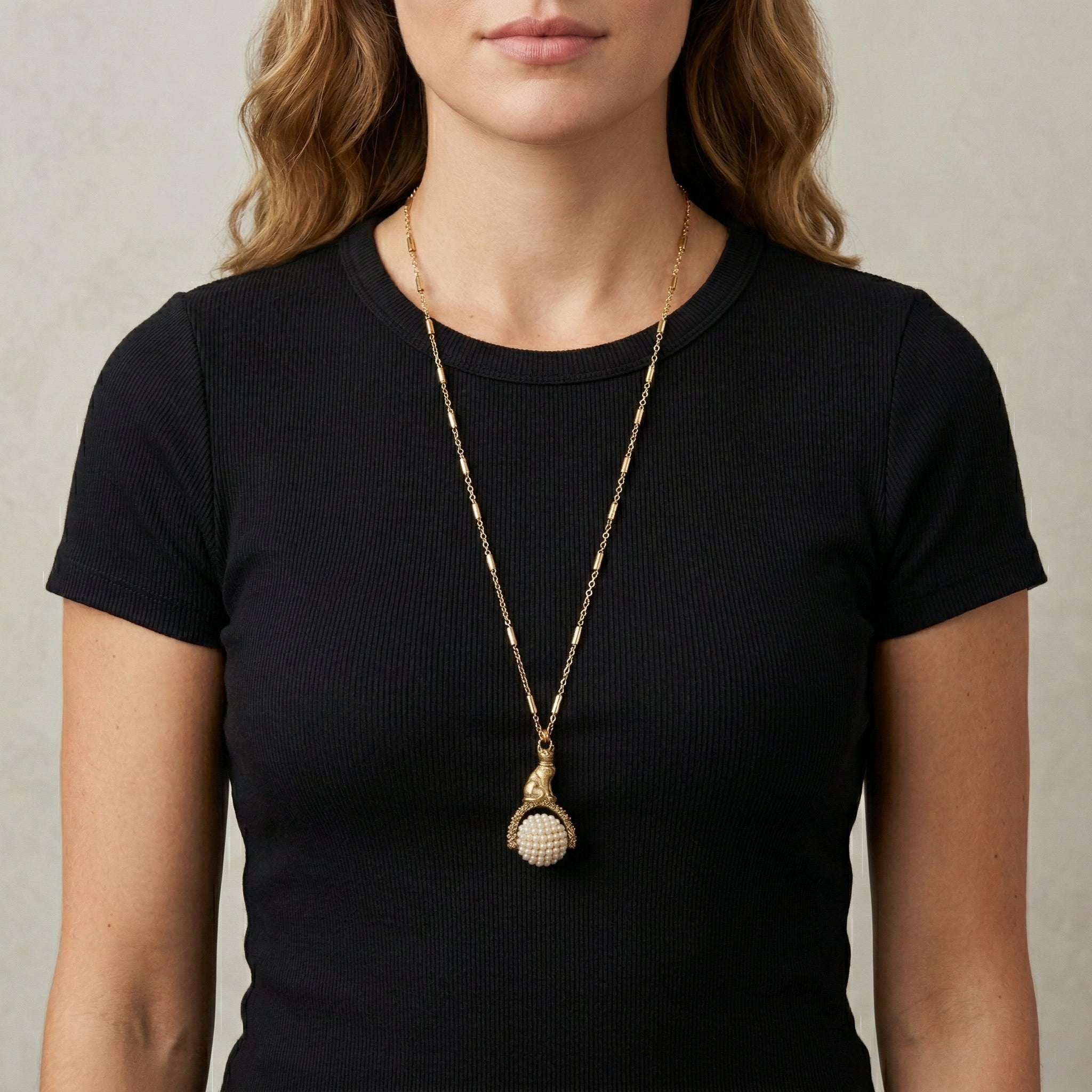 Woman wearing a black top with a gold tone cat pendant spinner necklace that has white faux pearls
