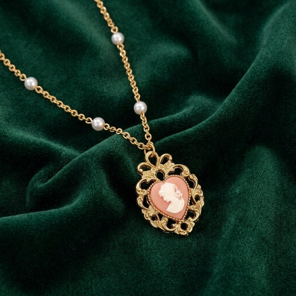 Gold tone chain necklace with white faux pearls and a pink heart-shaped cameo pendant resting on dark green fabric 