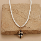 White faux pearl necklace with black stone cross pendant and crystal accents resting on light-colored sandstone