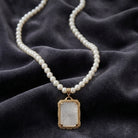 Pearl necklace with a gold tone framed pendant of glass and a clear crystal center on a dark fabric background
