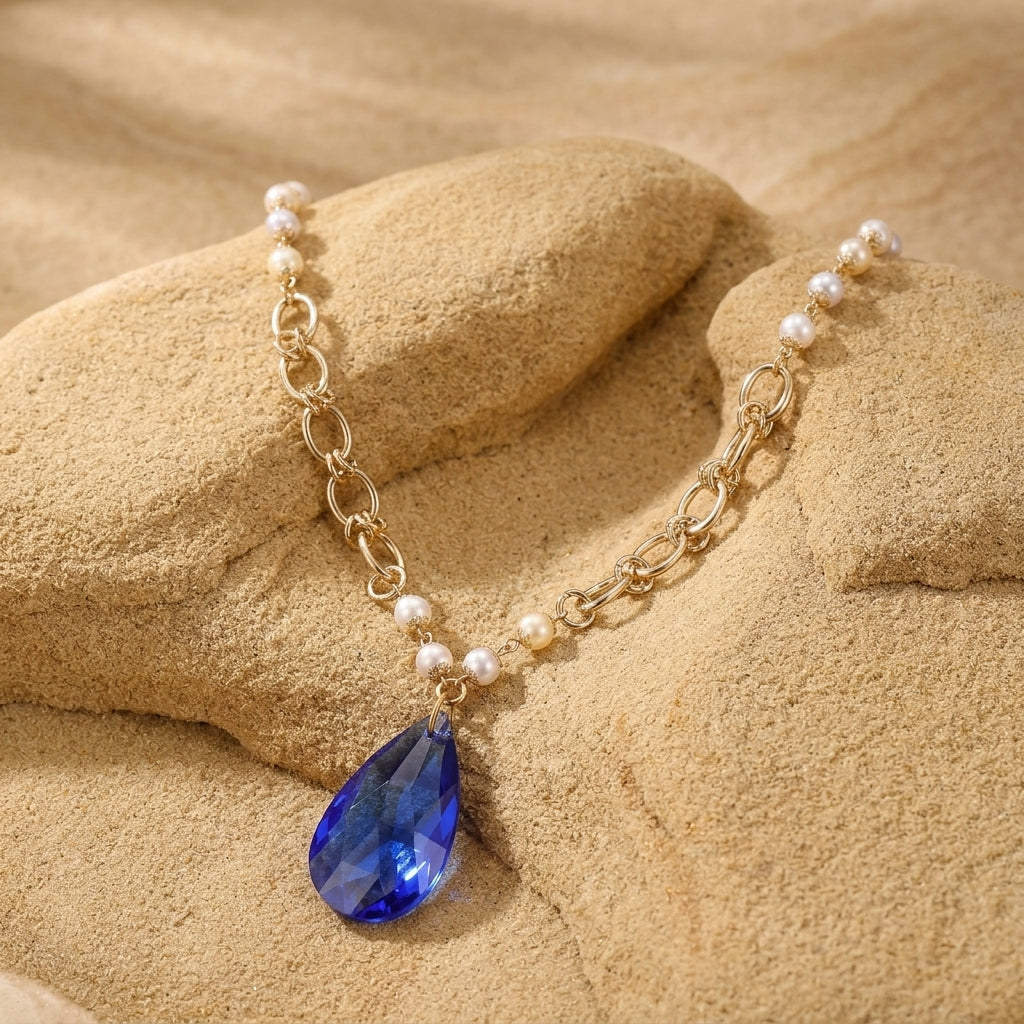 Gold tone link chain with faux pearls and a blue crystal pendant necklace resting on light stone surface