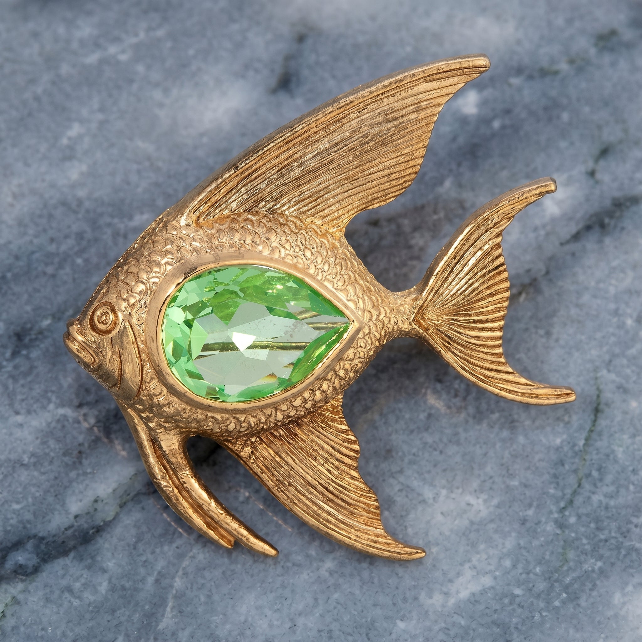 Gold fish-shaped brooch with a green crystal on a gray stone background