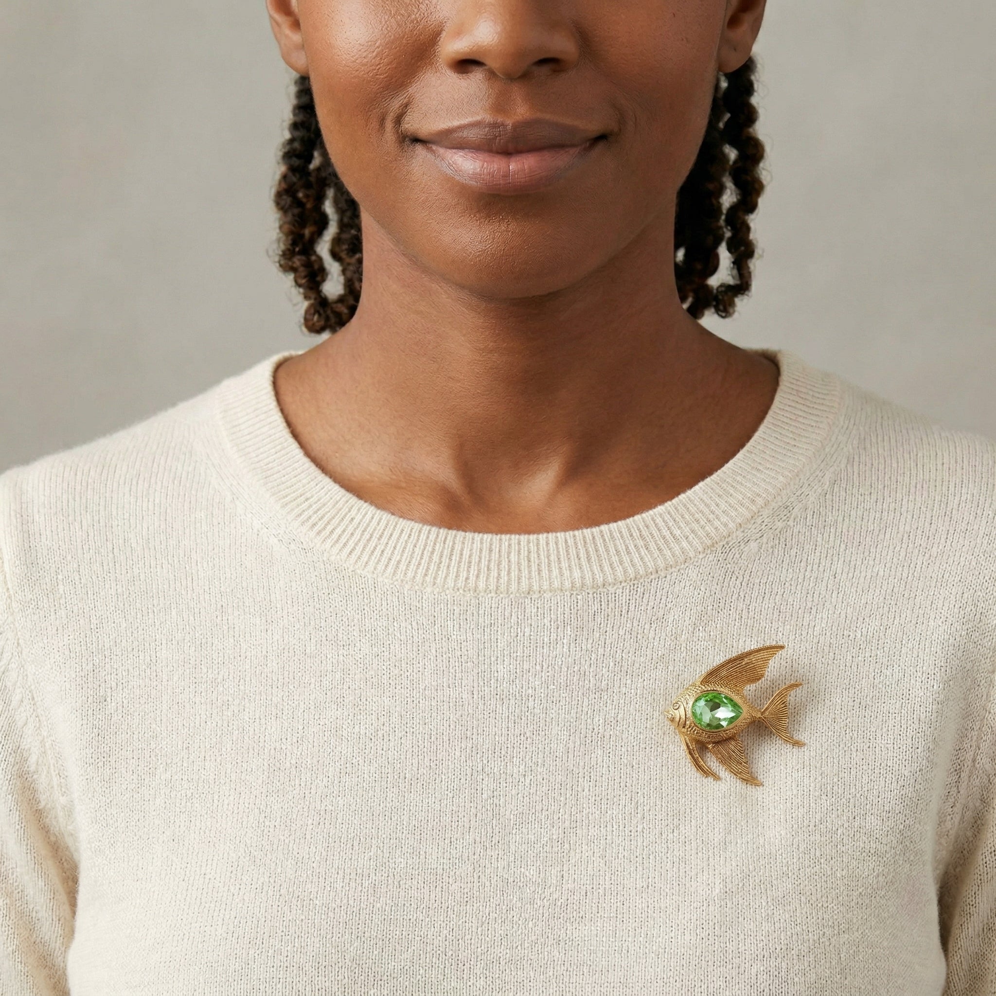 Woman wearing a beige sweater with a decorative angelfish brooch with a green crystal center on a neutral background