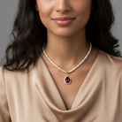 Woman wearing a faux pearl necklace with a red gemstone teardrop pendant lined by crystals against a neutral background