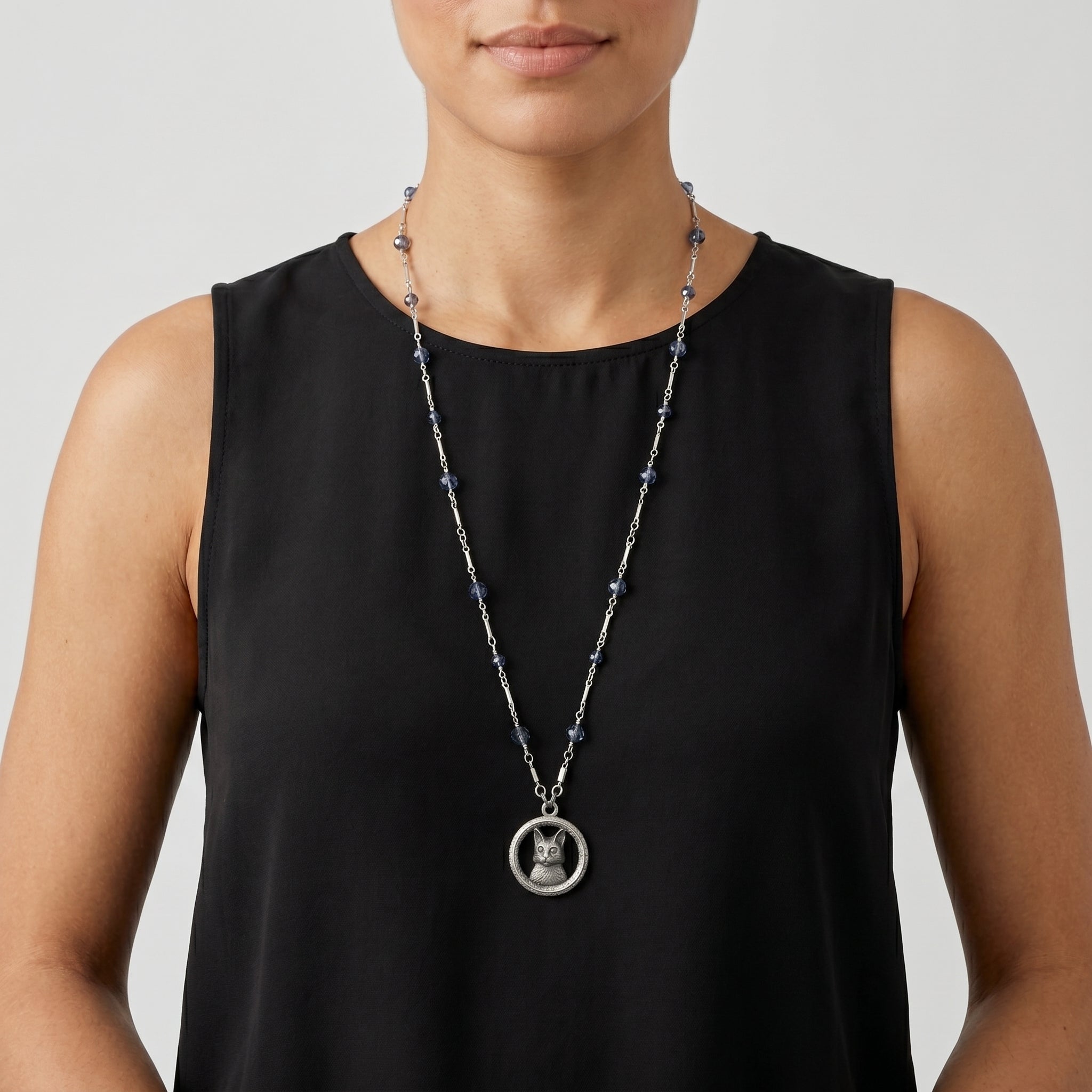 Person wearing a long silver necklace that has blue crystal beads with a circular cat pendant on a plain background