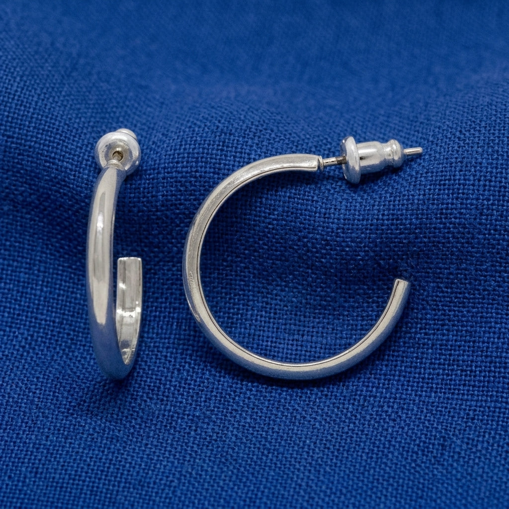 A pair of polished silver hoop earrings shown from different angles on a textured blue fabric background.