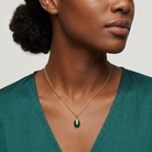 Woman wearing a gold necklace with a green egg-shaped pendant with clear crystal accents against a neutral background