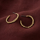 Twisted gold hoop earrings with post backs, displayed on a textured dark burgundy fabric background.