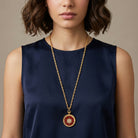 Woman wearing a gold necklace with a red starburst pendant on a neutral background