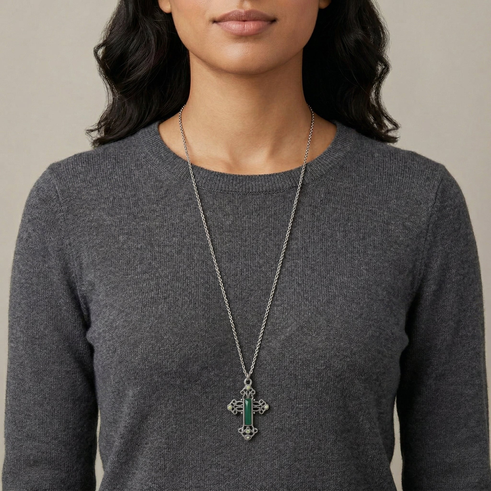 Person wearing a long necklace with a cross pendant and green crystals and gemstones on a neutral background
