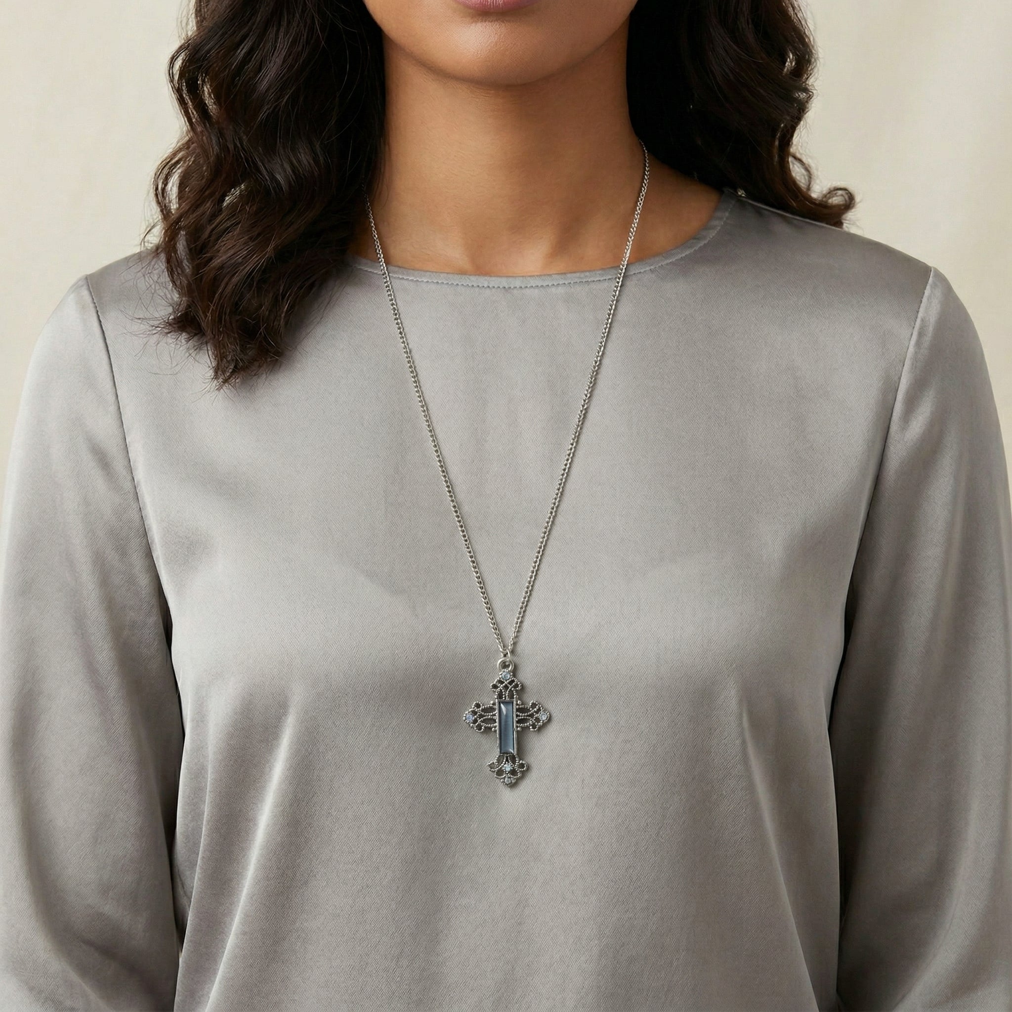 Person wearing a long-sleeve gray top with a necklace featuring a cross pendant and blue crystals and gemstones.