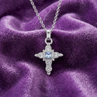 Silver cross pendant with a blue crystal center and clear crystal accents on a purple fabric background