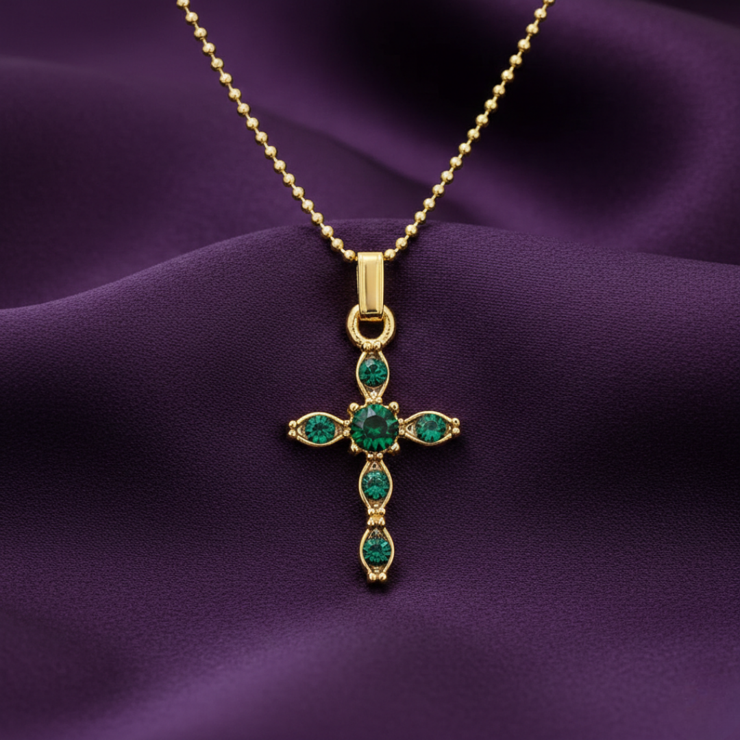 Gold cross pendant with green gemstones on a purple fabric background