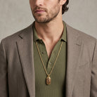 Man wearing a brown blazer and green shirt with a gold pendant necklace against a neutral background