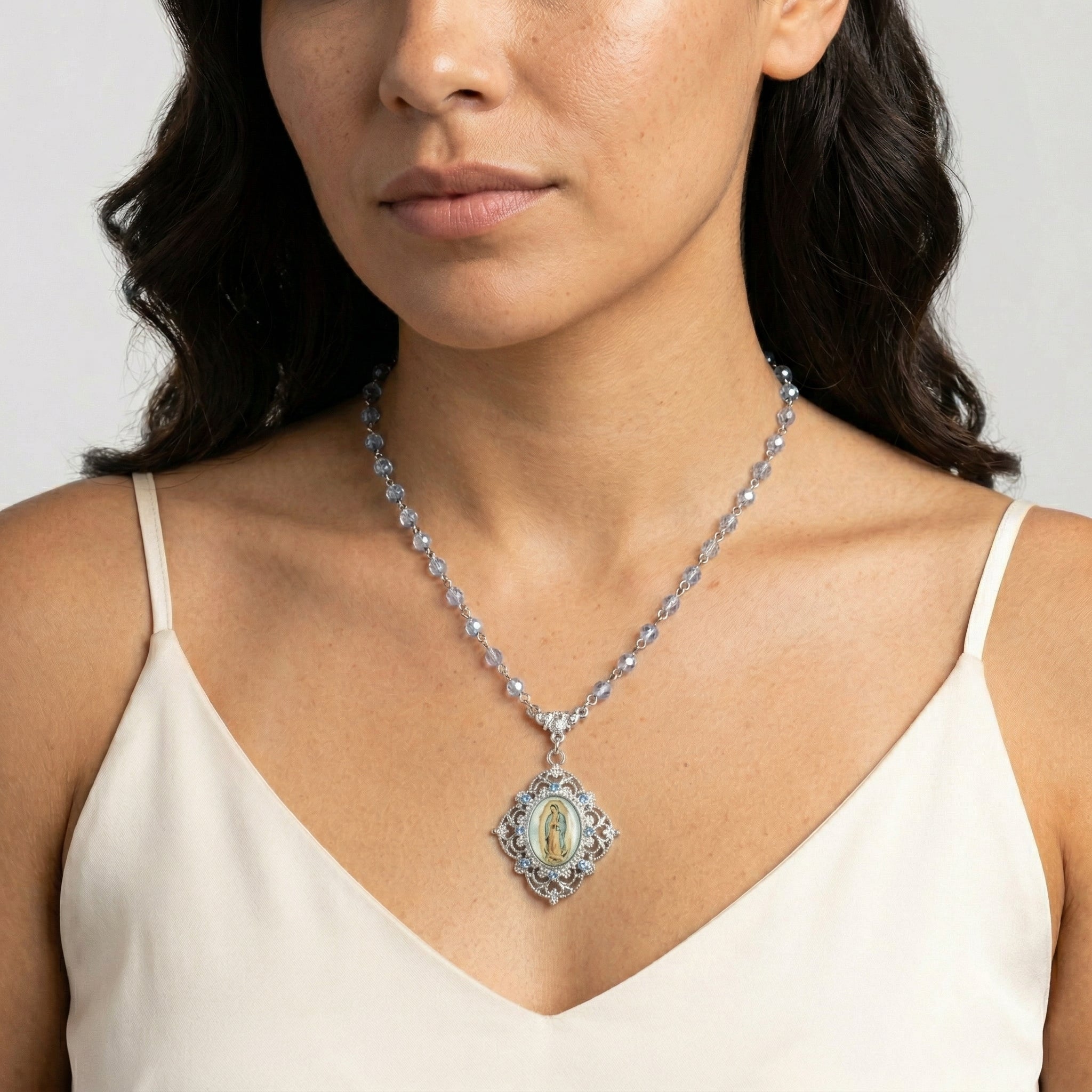 Woman wearing a silver tone necklace with crystals on the chain and an Our Lady of Guadalupe pendant against a neutral background
