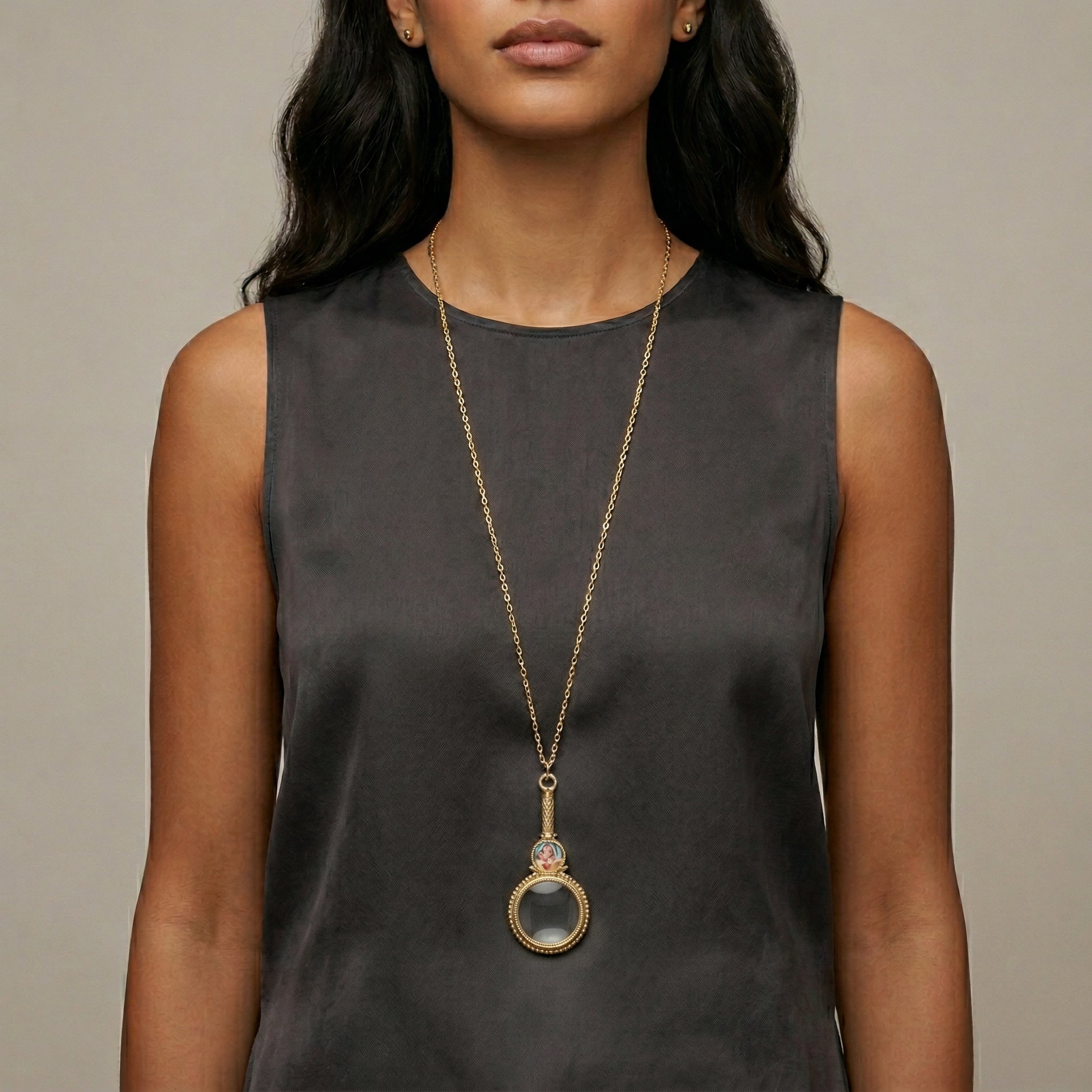 Woman wearing a long gold necklace with a magnifying glass pendant and Mother Mary and Child decal against a neutral background