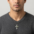 Man wearing a gray sweater with a silver cross necklace featuring a green gemstone on a neutral background