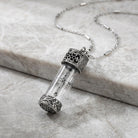 Silver necklace with a clear glass vial pendant and cross design on a marble surface