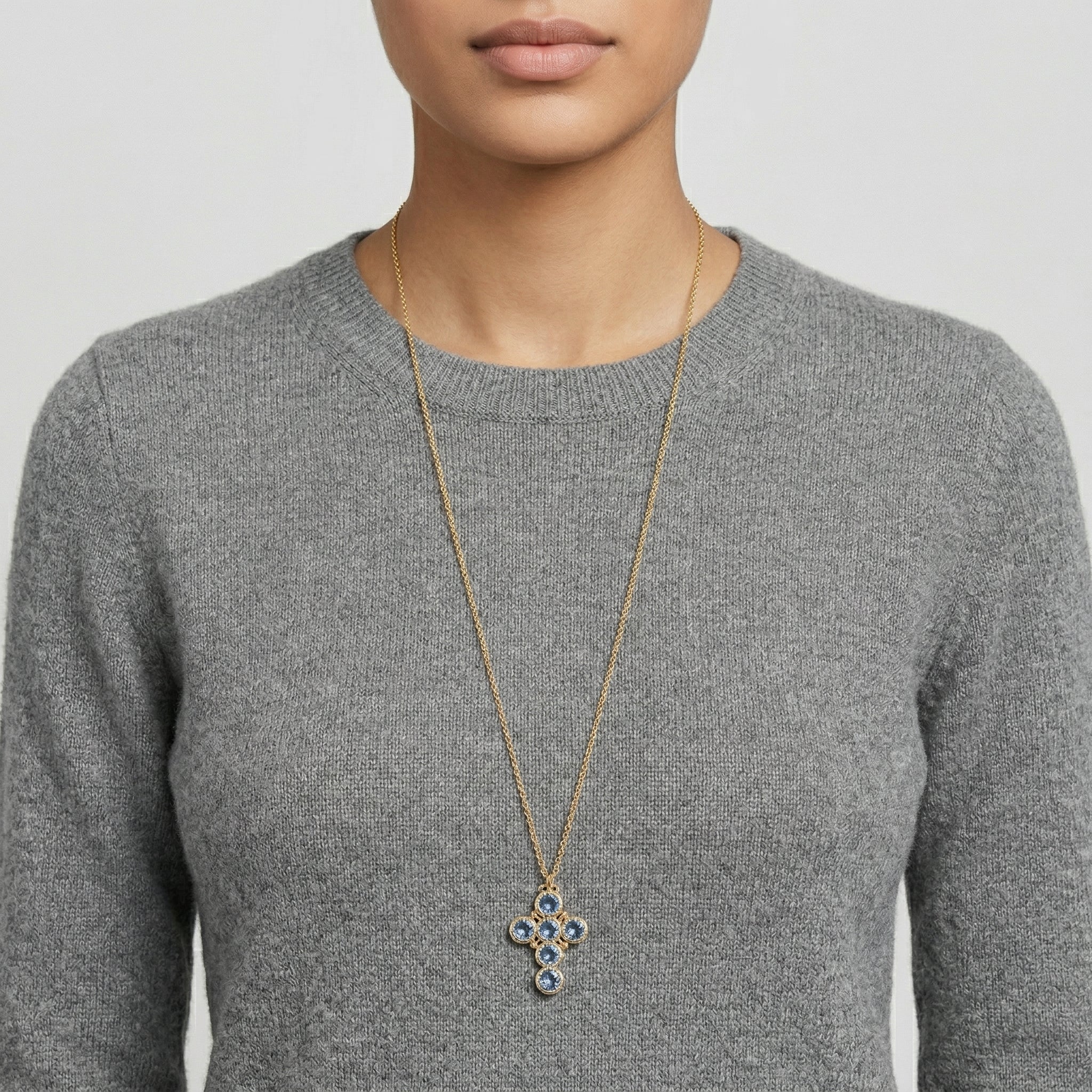Person wearing a long necklace with a blue crystal cross pendant on a plain background