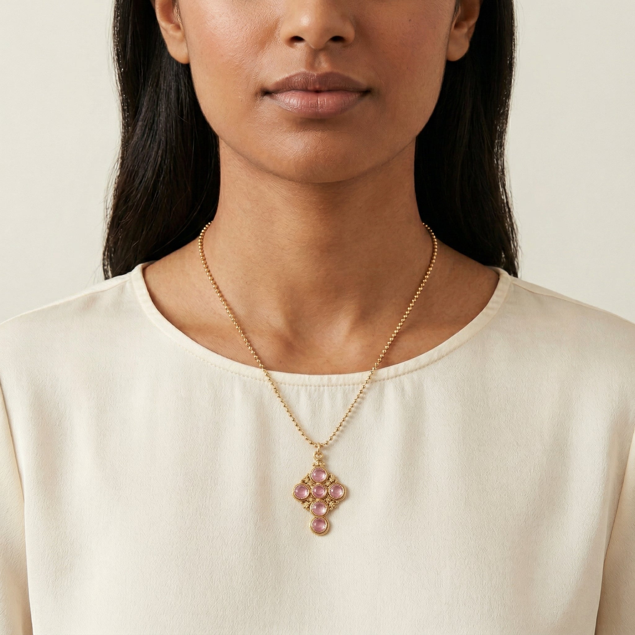 Woman wearing a gold necklace with a pink crystal and cross pendant against a neutral background