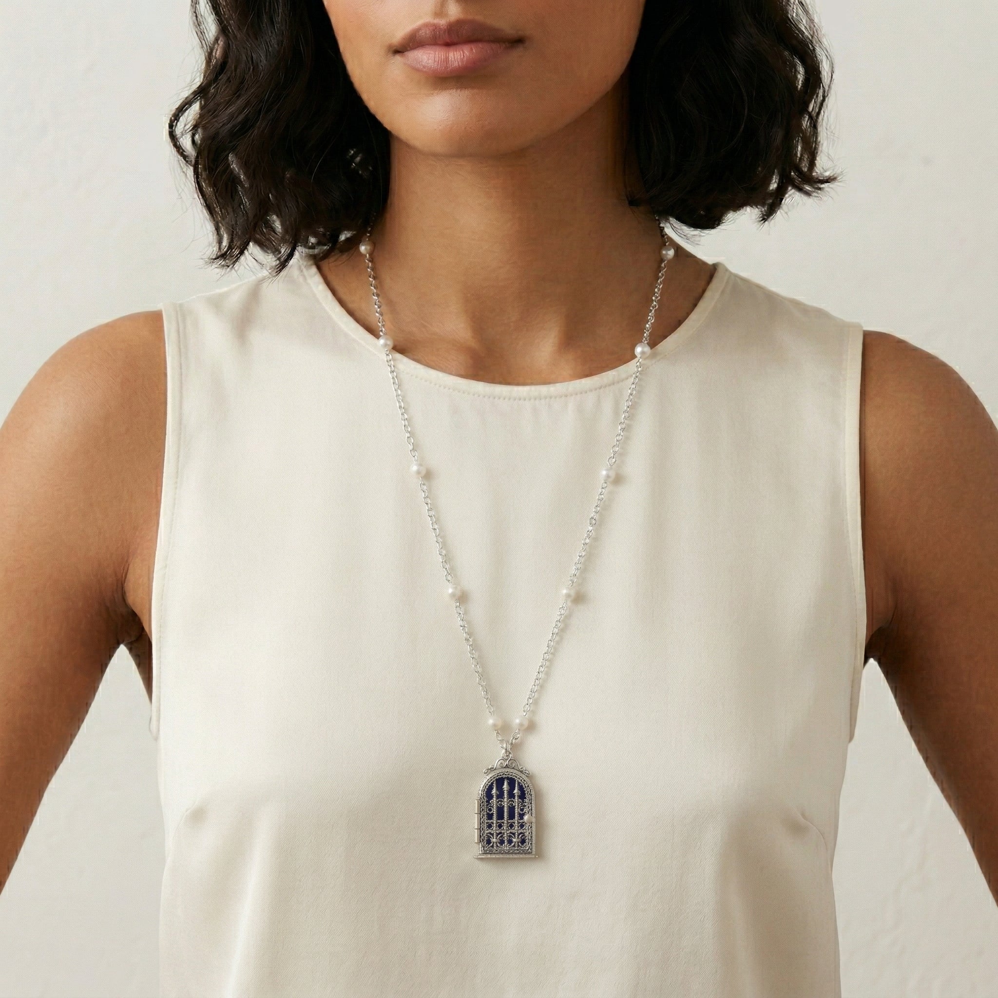 Woman wearing a silver necklace with faux pearls and a cross locket pendant against a neutral background
