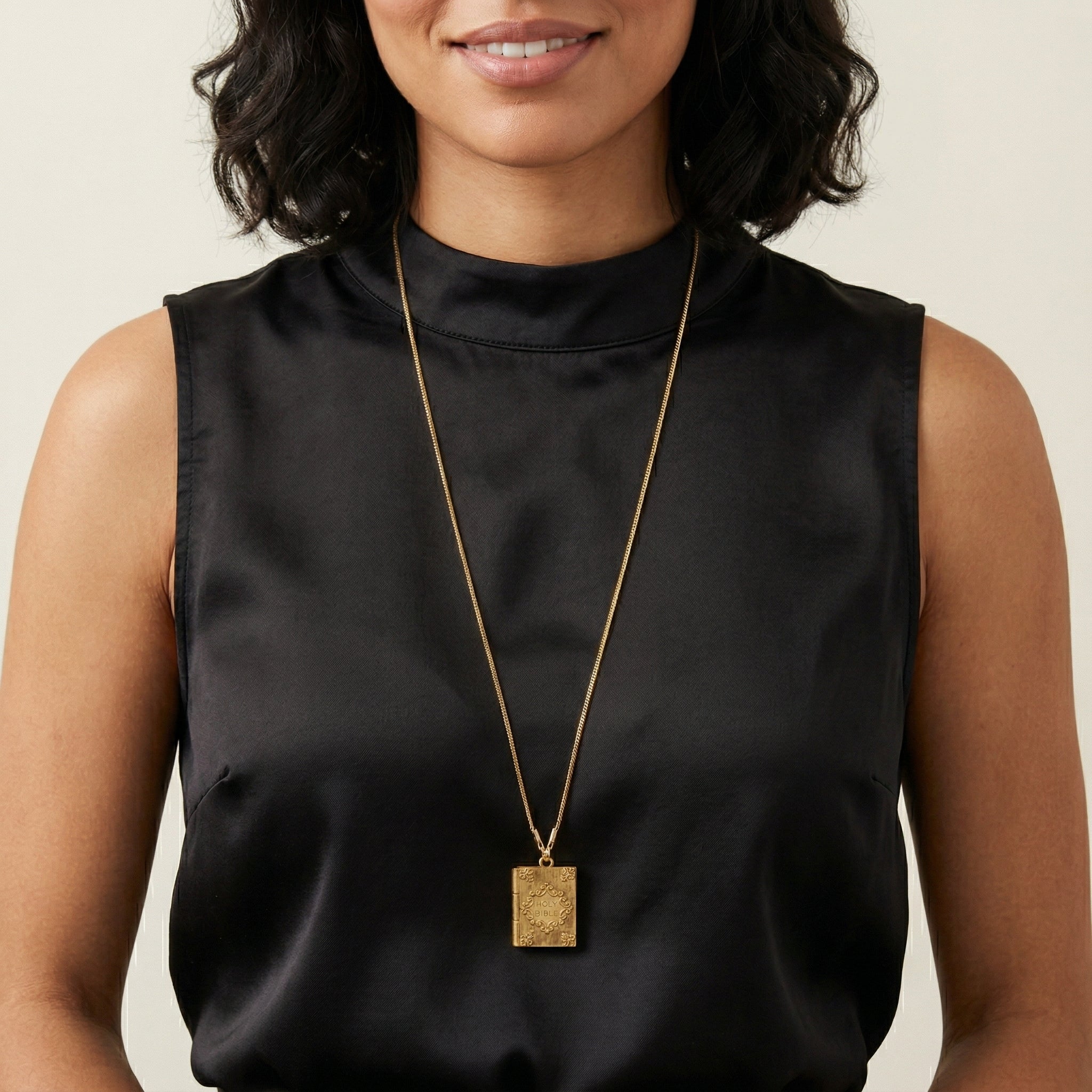 Woman wearing a black sleeveless top and gold necklace with a square Holy Bible pendant.